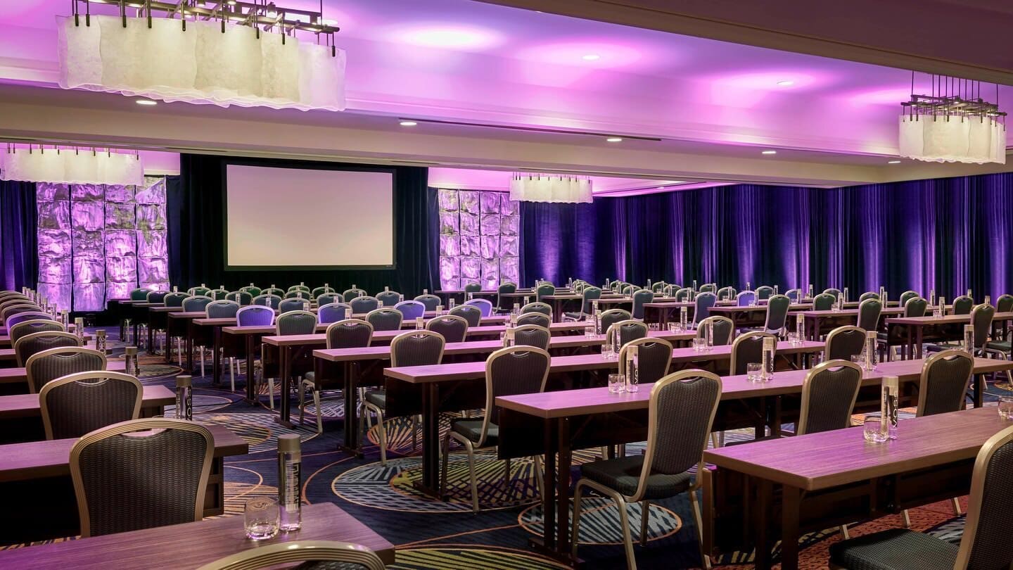 A large conference room set up with rows of tables and chairs facing a projection screen, illuminated by overhead lights and purple accent lighting along the walls.