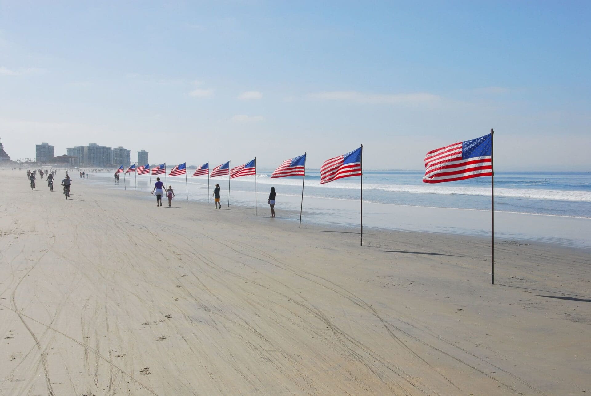 Rows of American flags lined up along a sandy beach with people walking nearby. The ocean waves and buildings are visible in the background under a blue sky.