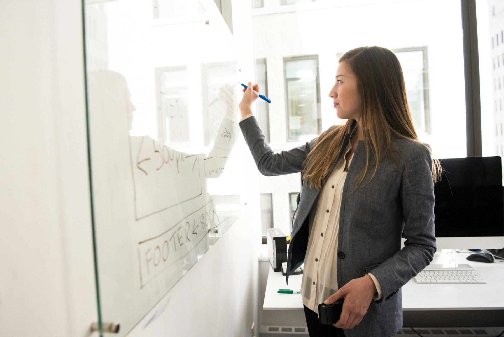 A woman in business attire writes on a whiteboard in a modern office, holding a marker and a phone, with diagrams and notes visible on the board. A computer and office supplies are on the desk behind her.