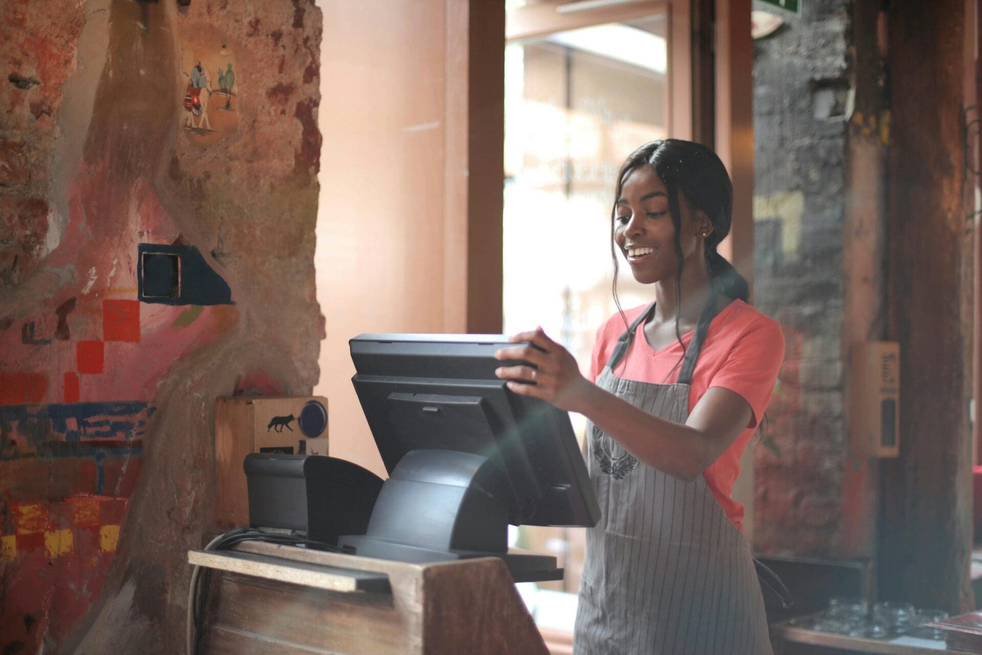 A woman wearing an apron stands behind a counter, smiling as she uses a touchscreen register in a warmly lit, rustic cafe or restaurant.
