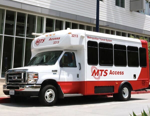 A white and red Metropolitan Transit System (MTS) Access shuttle bus is parked on a city street in front of a modern building. The bus has large windows and the MTS Access logo on its side.