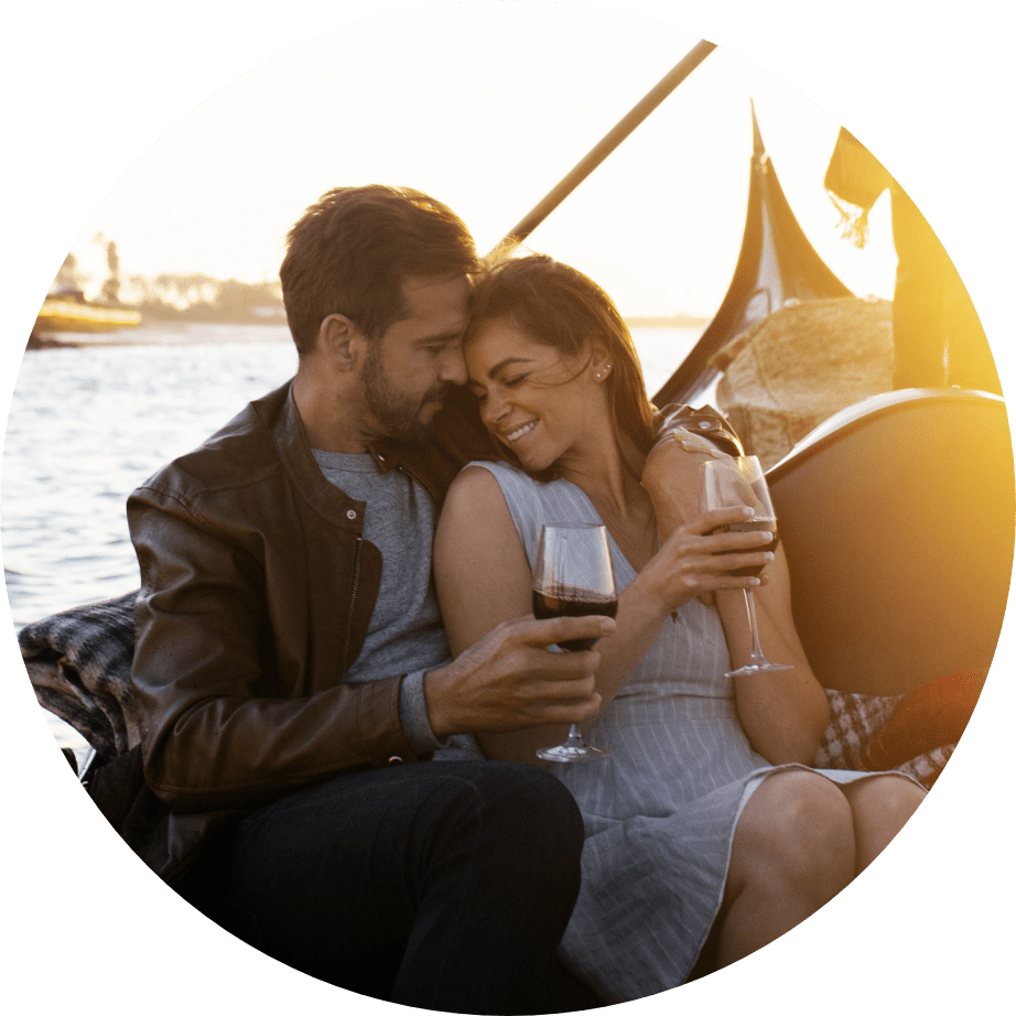A couple sits close together on a boat at sunset, smiling and holding glasses of red wine, with water and a gondola in the background.