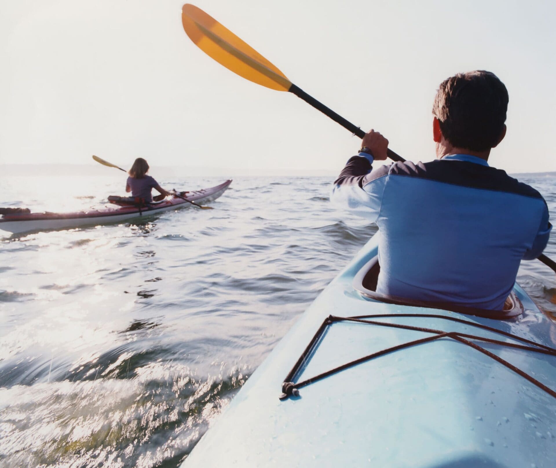Two people kayaking on open water under bright sunlight, with one person in the foreground and another further ahead. The water is gently rippling and the sky is clear.